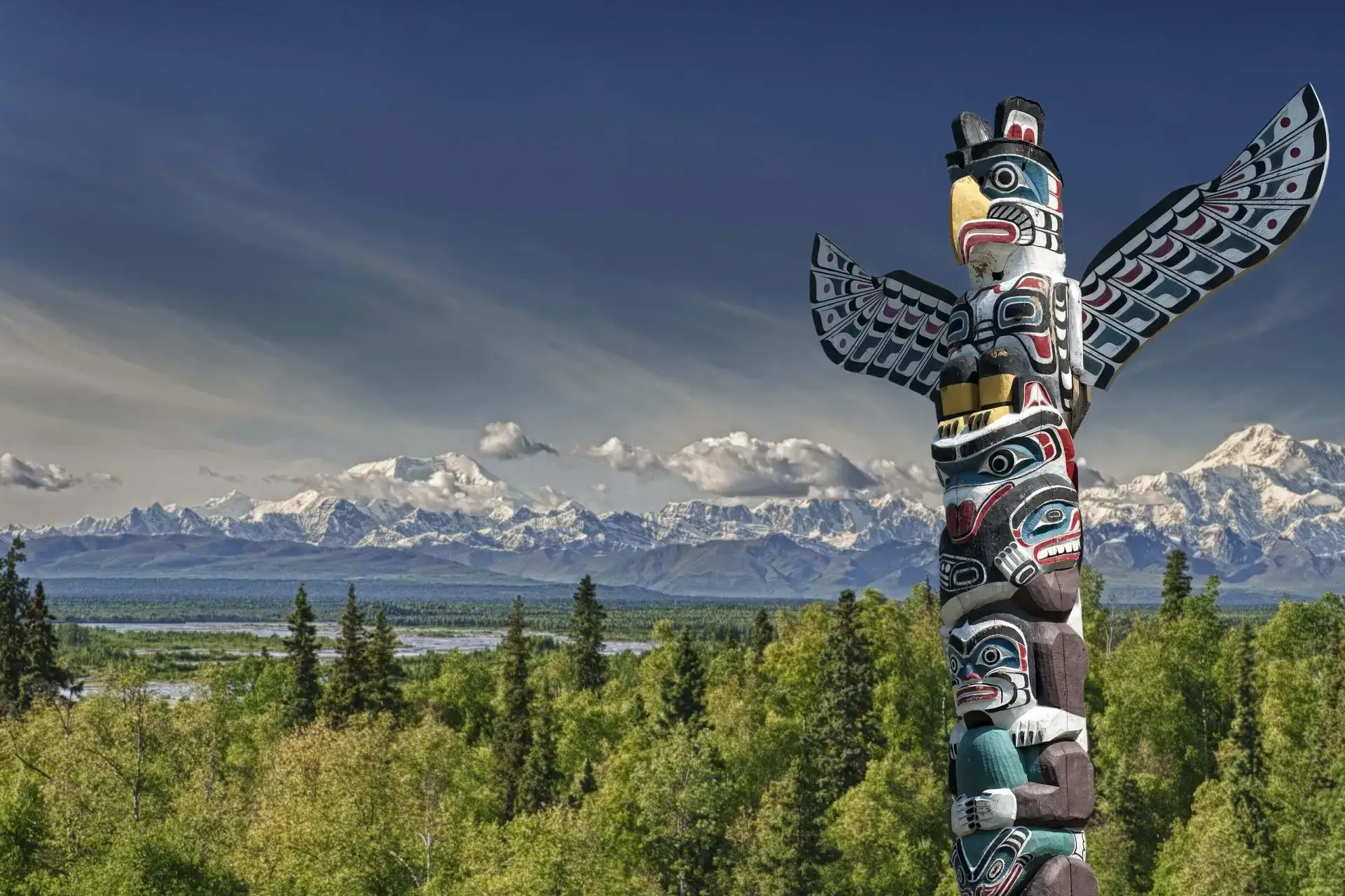 A colorful totem pole stands against a scenic mountain and forest backdrop.